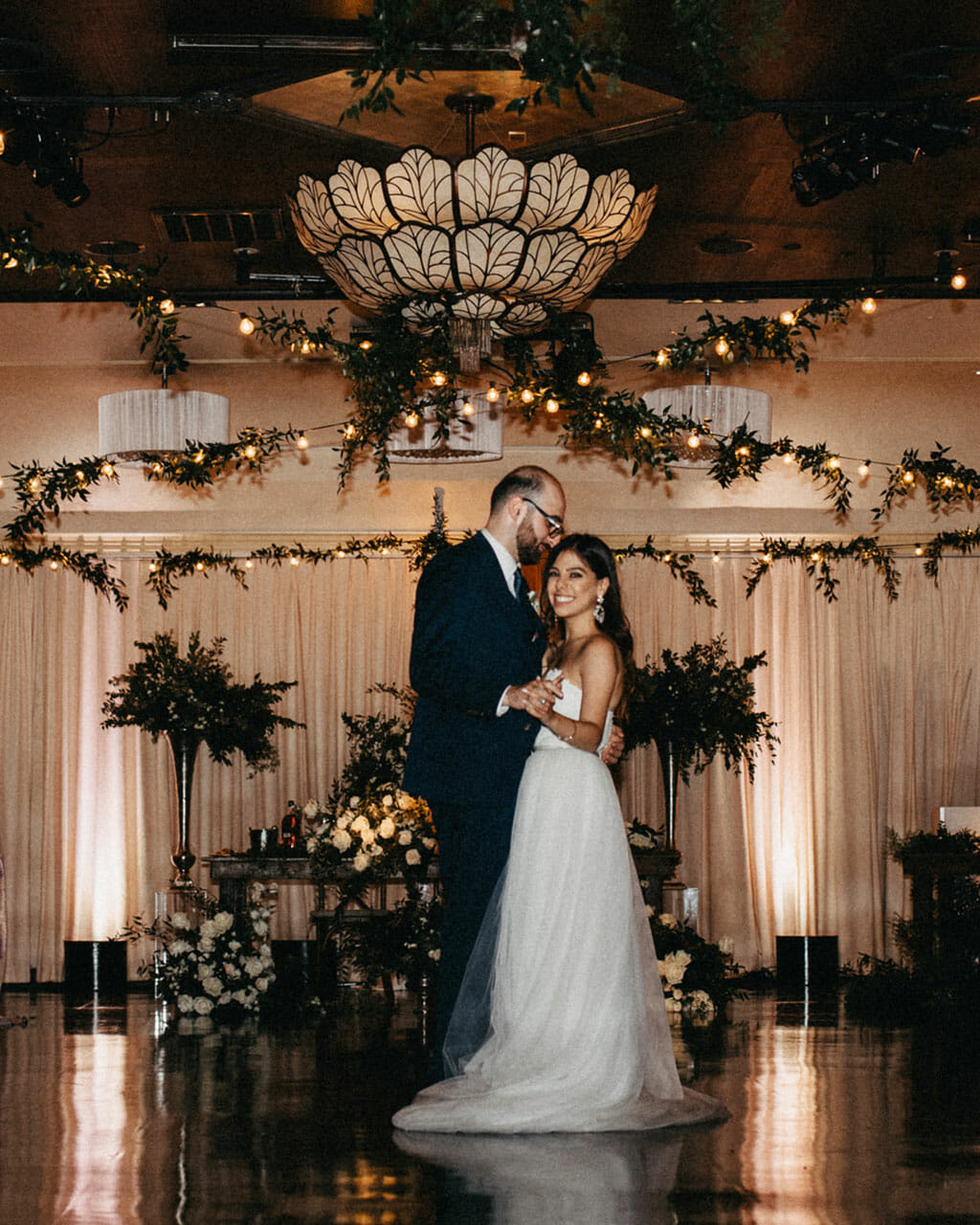 wedding couple standing under noor's sofia banquet hall chandelier with greenery wedding reception decor and string lights