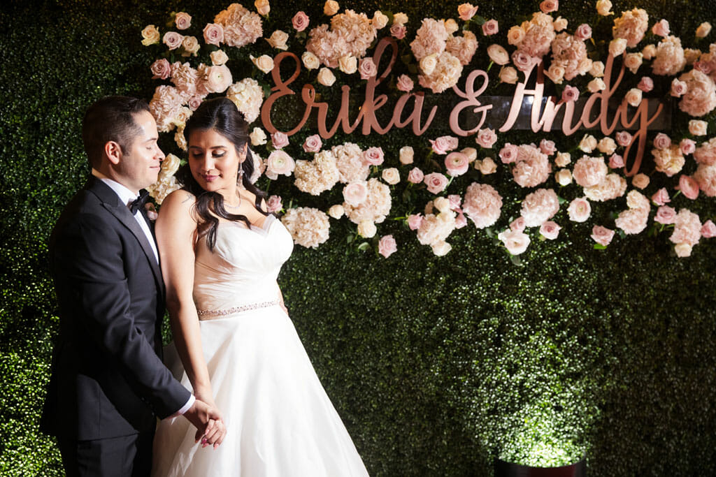 bride and groom standing in front of flower wall with green grass backdrop and pink flowers and their names in gold