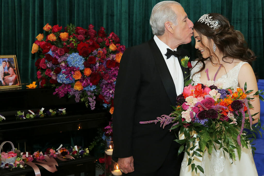 Father of the bride kissing his daughters head with flowers behind them