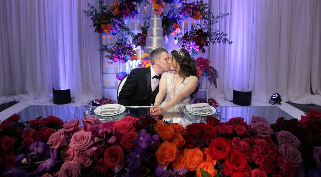 bride and groom seated at sweetheart table surrounded by red orange and pink flowers and a wedding cake behind them