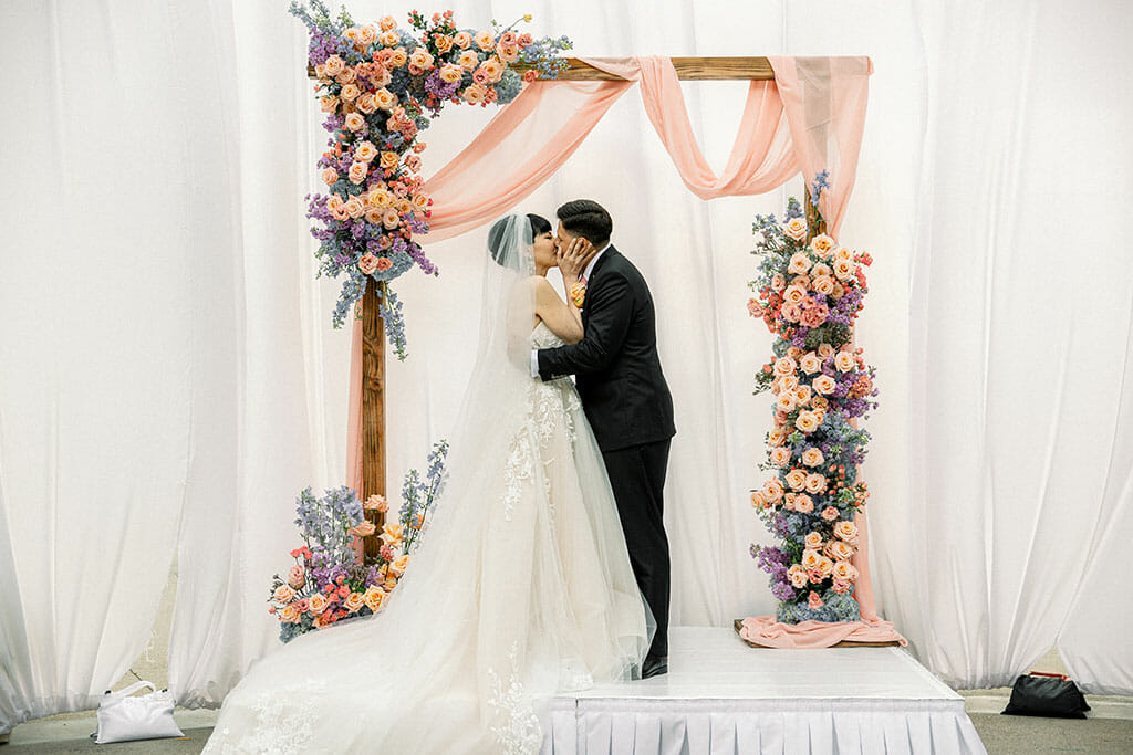NOOR los angeles wedding ceremony on the NOOR terrace with bride and groom kissing under a pink and pastel wedding arch against a white satin backdrop